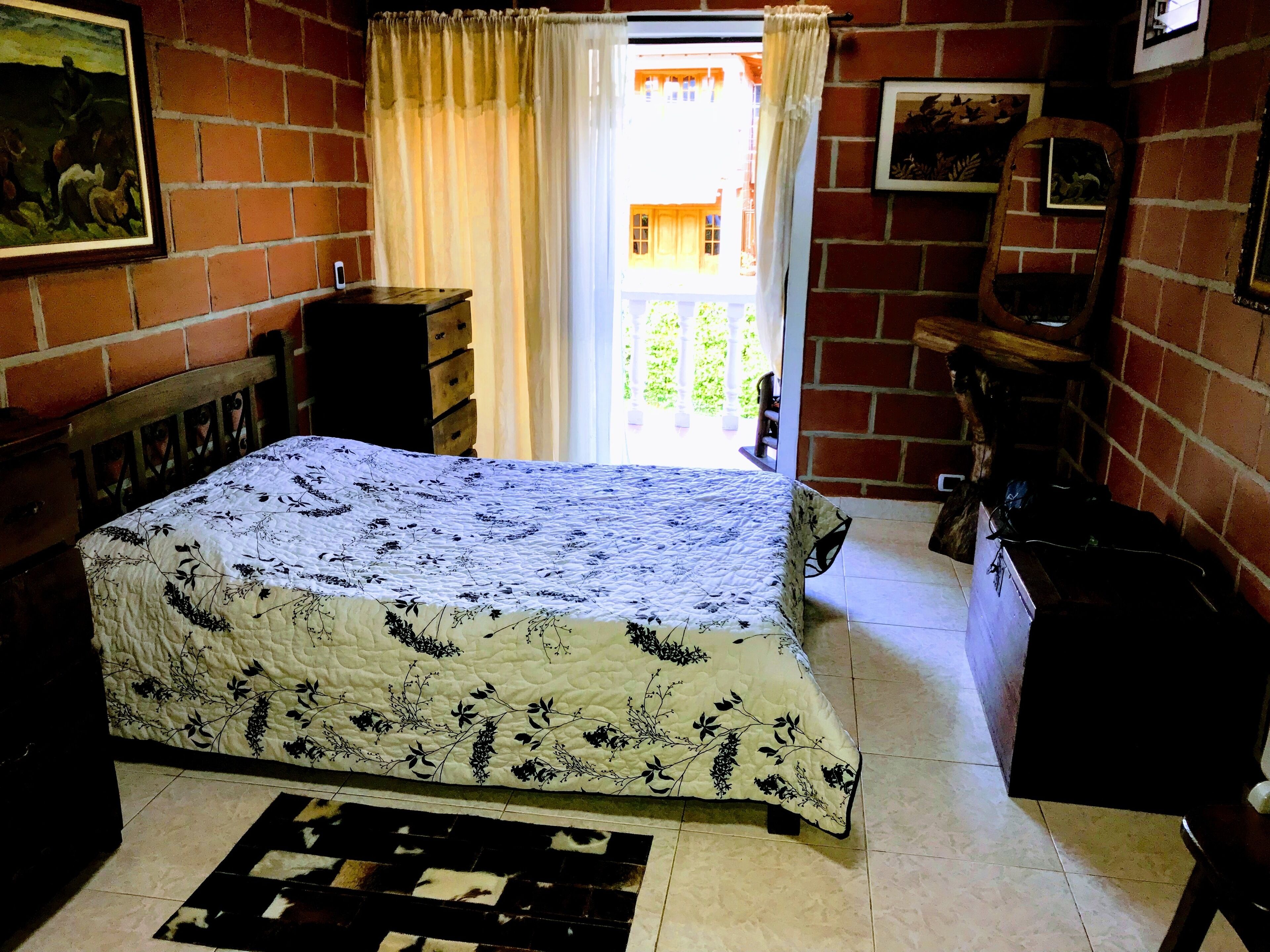 Une chambre à coucher avec un lit, une commode, une fenêtre avec des rideaux et un mur en briques.
