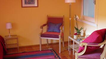 A room with a wooden floor, a red and blue striped rug, a wooden chair with a cushion, a small table with a lamp, and a window with a view of the outside.
