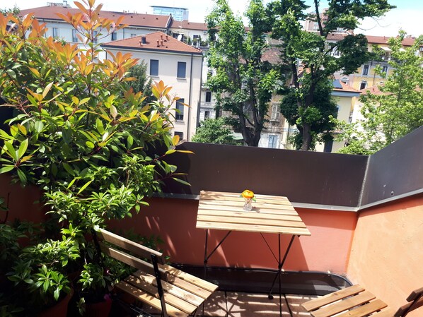 A wooden table with a vase of yellow flowers, a bench, and a view of buildings and trees.
