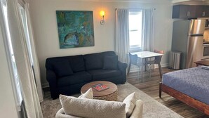 A modern hotel room with a dark-colored sofa, a small round table, a bed with a blue quilt, a painting on the wall, and a kitchenette with a refrigerator and microwave.