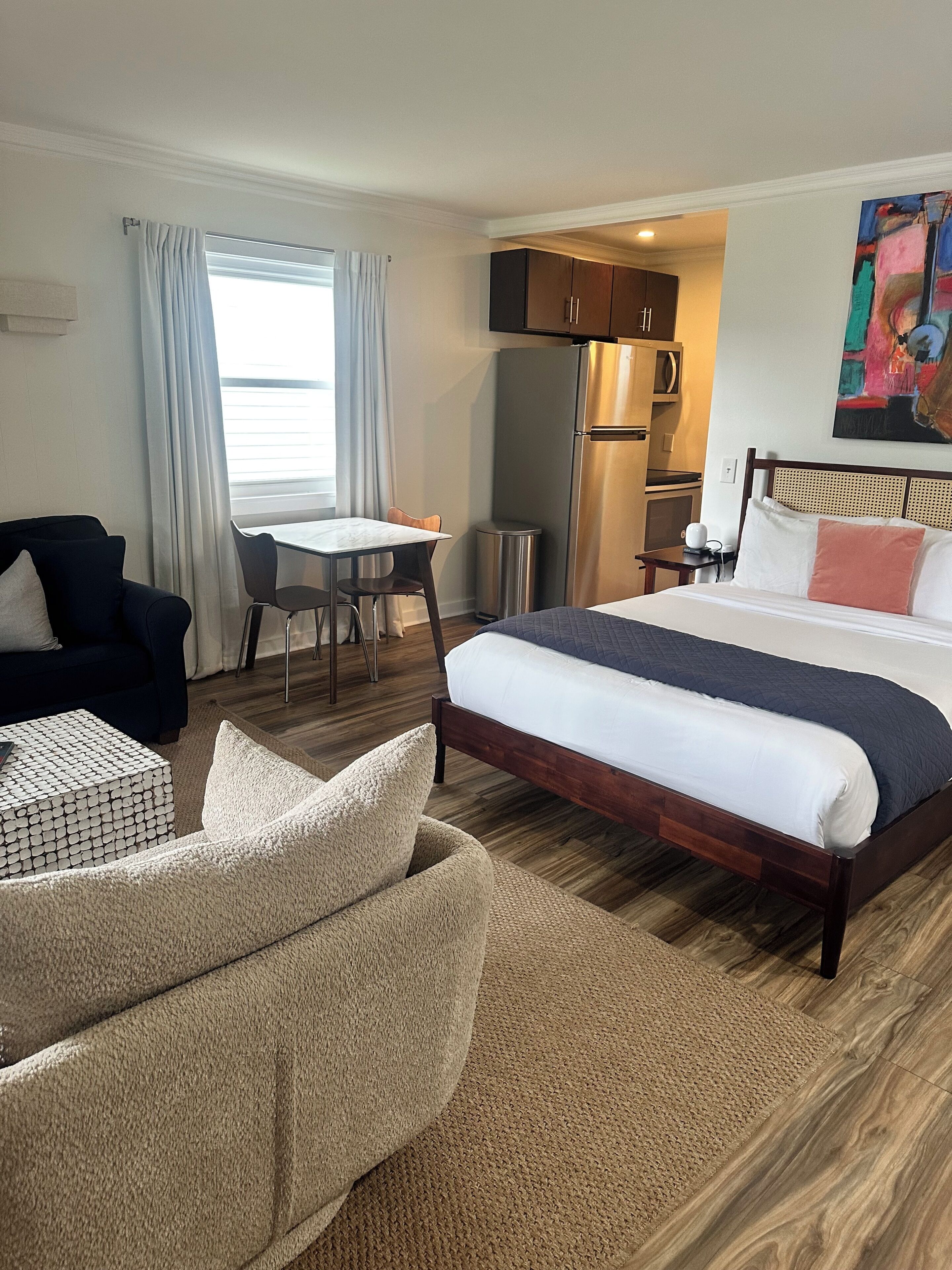 A hotel room with a bed, a desk, a chair, a sofa, a trash bin, and a painting on the wall.