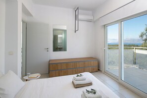 Een moderne slaapkamer met een groot bed, een houten dressoir, een spiegel, en een balkon met uitzicht op zee en bergen.