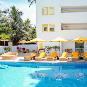 A pool area with lounge chairs and umbrellas, a white building with yellow-framed windows, and palm trees in the background. - Bloomrooms @ Calangute (Calangute)
