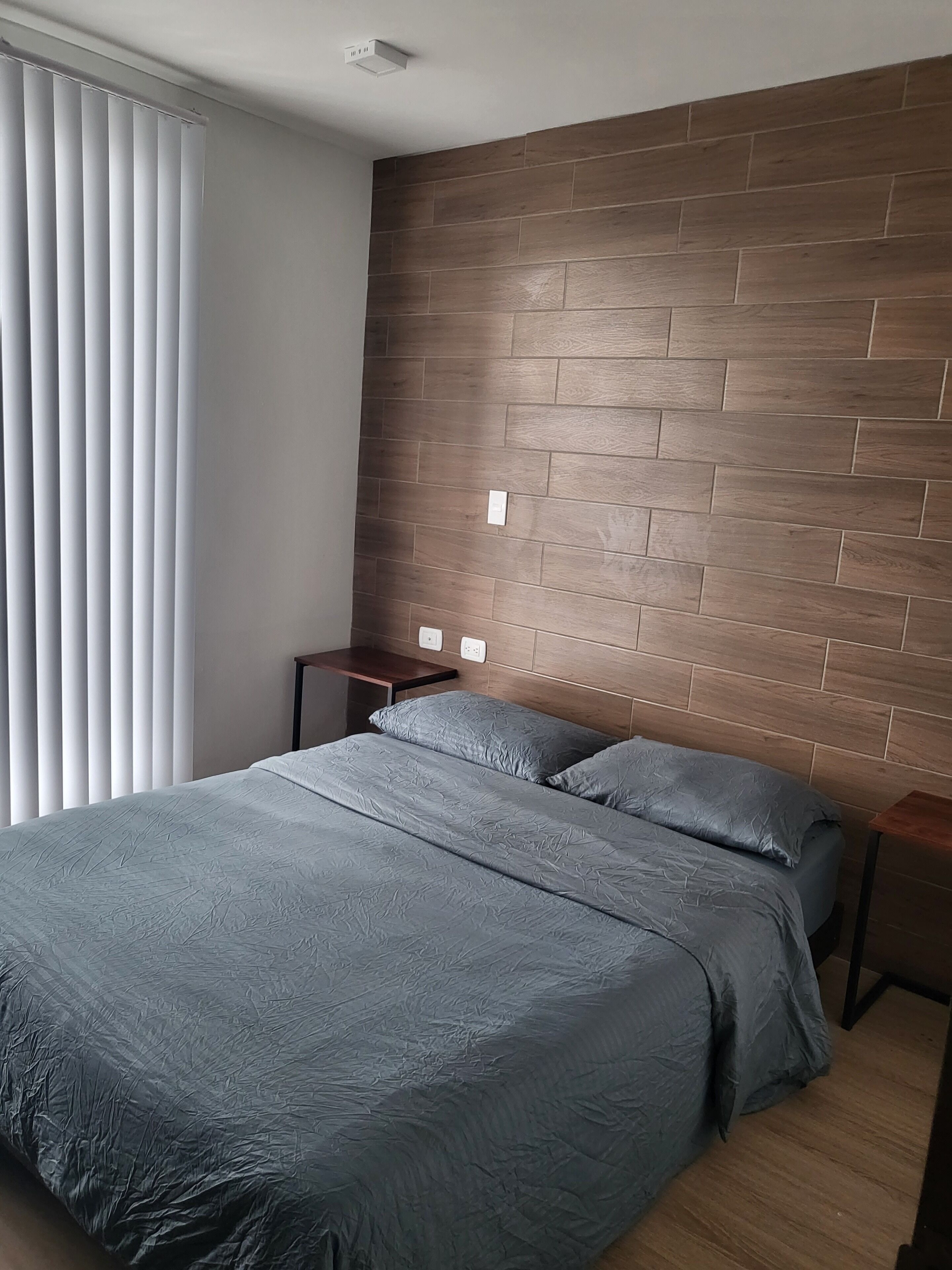 Un dormitorio con cabecera de madera, una cama con ropa de cama gris, una mesita de noche y una ventana con persianas.