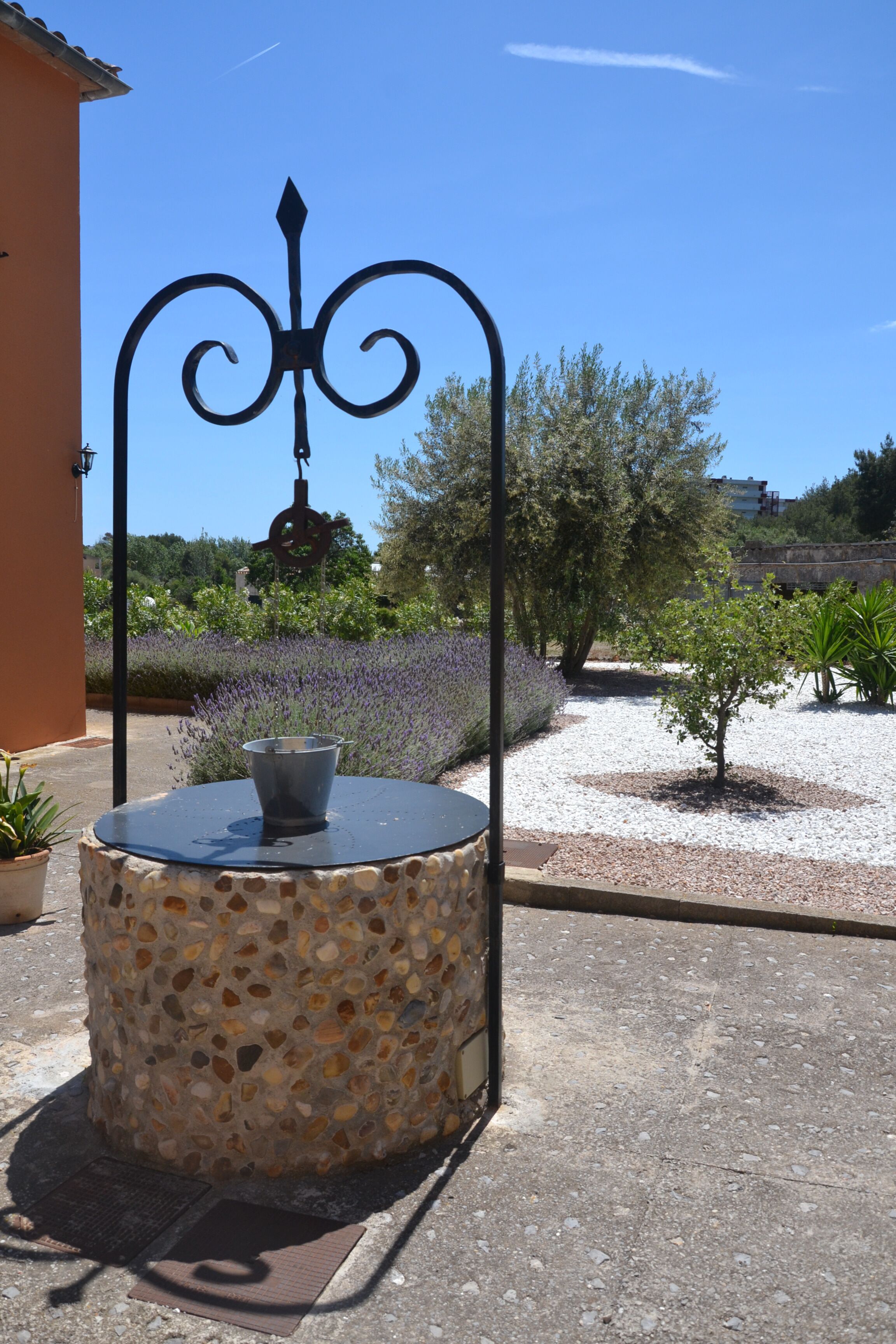 A round stone table with a metal frame and a metal bowl, set on a paved area with a clear blue sky and trees in the background.