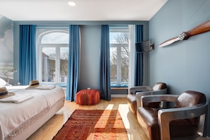 A hotel room with a bed, a red ottoman, leather chairs, a TV mounted on the wall, and a large window with curtains. - Casa da Praça (Porto)