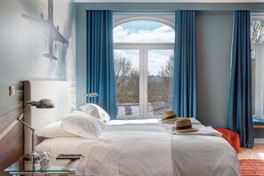 A hotel room with a large bed, a bedside table with a lamp, a window with curtains, and a view of the outside. - Casa da Praça (Porto)
