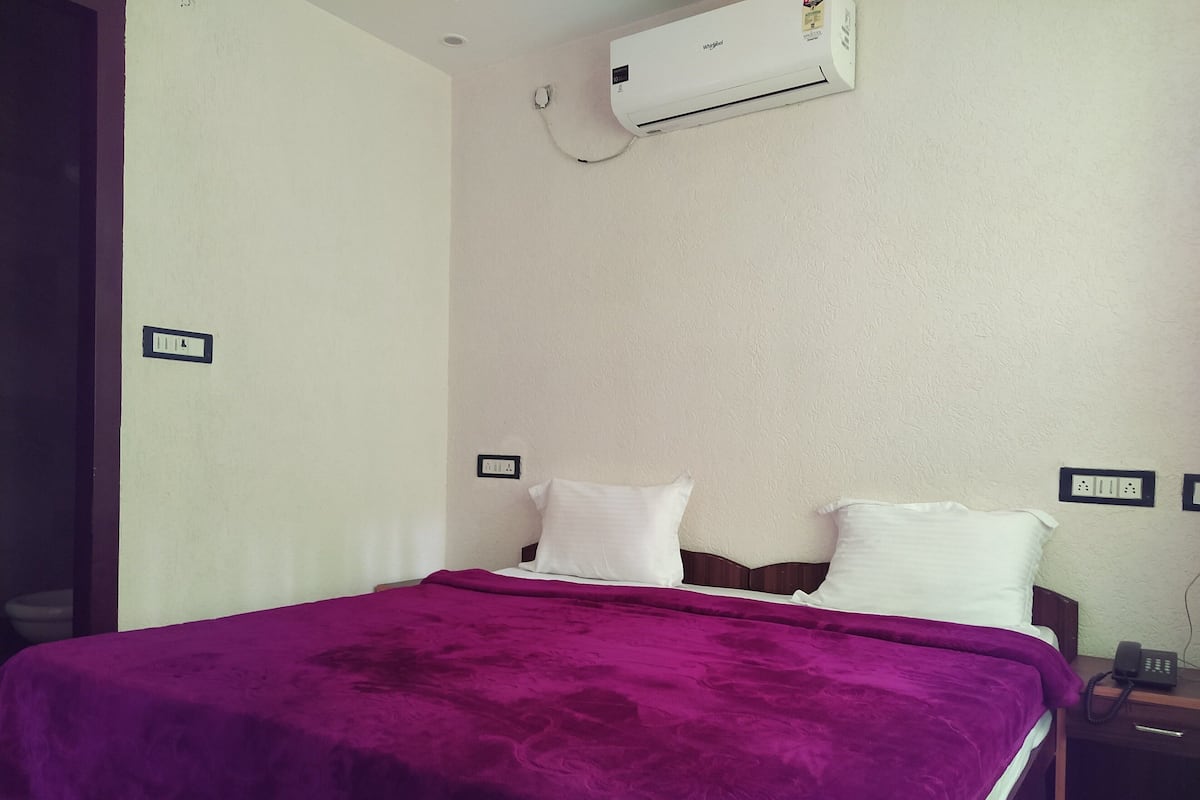 A bedroom with a bed, a purple blanket, white pillows, a nightstand with a phone, and an air conditioner on the wall.