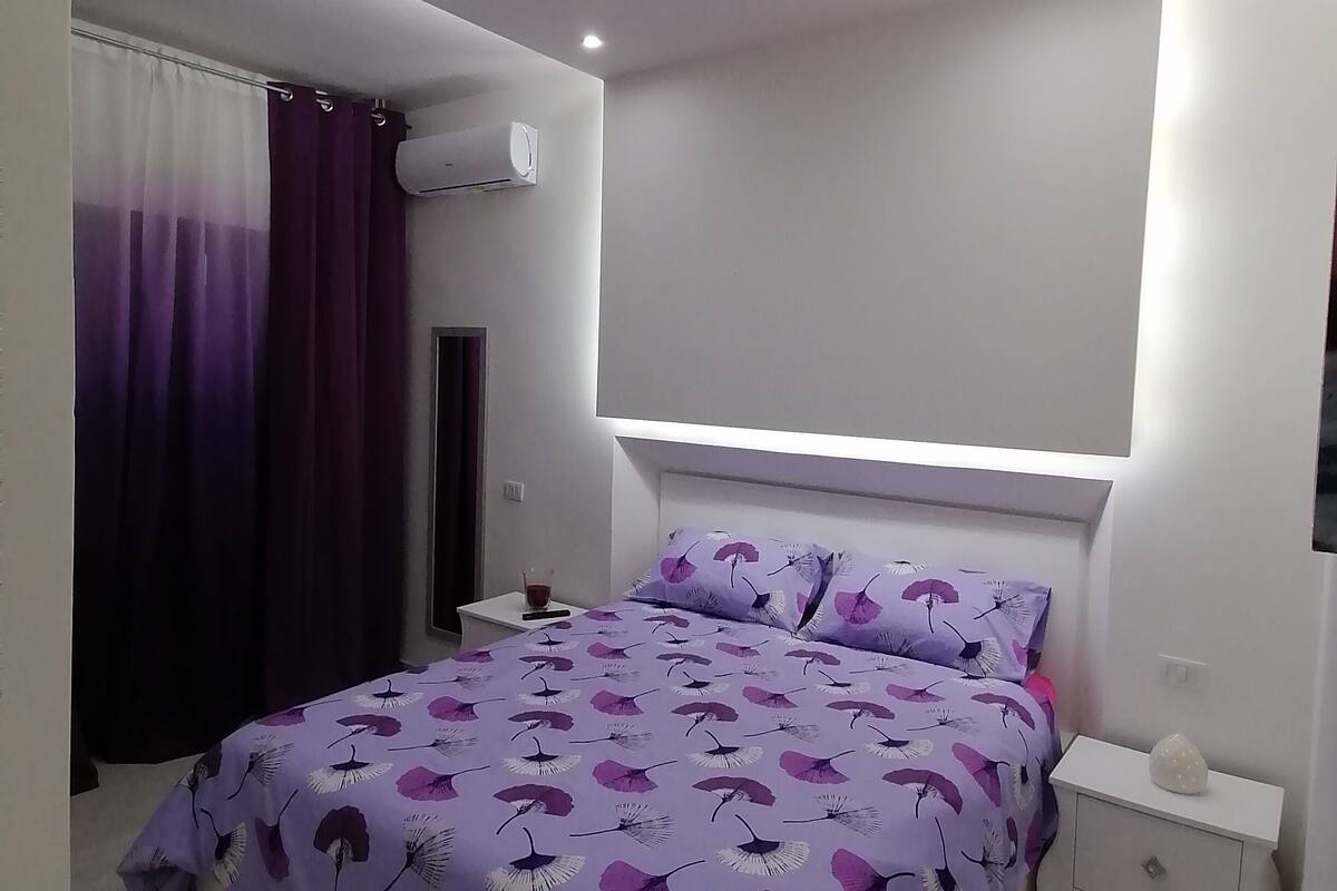 Quarto moderno com cama, mesas de cabeceira, aparelho de ar condicionado e luminária no teto.