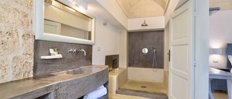 Un baño moderno con un espejo grande, un tocador de madera, zona de ducha y una puerta que da a otra habitación.