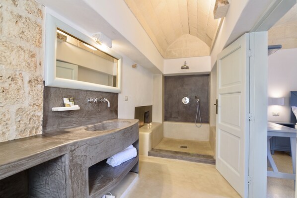 Un baño moderno con un espejo grande, un tocador de madera, zona de ducha y una puerta que da a otra habitación.