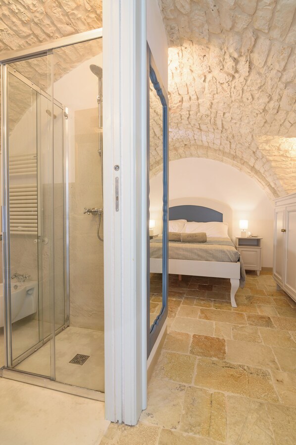A bedroom with a stone ceiling, a glass-enclosed shower, a bed with a blue headboard, and a nightstand with a lamp. - Masseria Ayroldi (Ostuni)