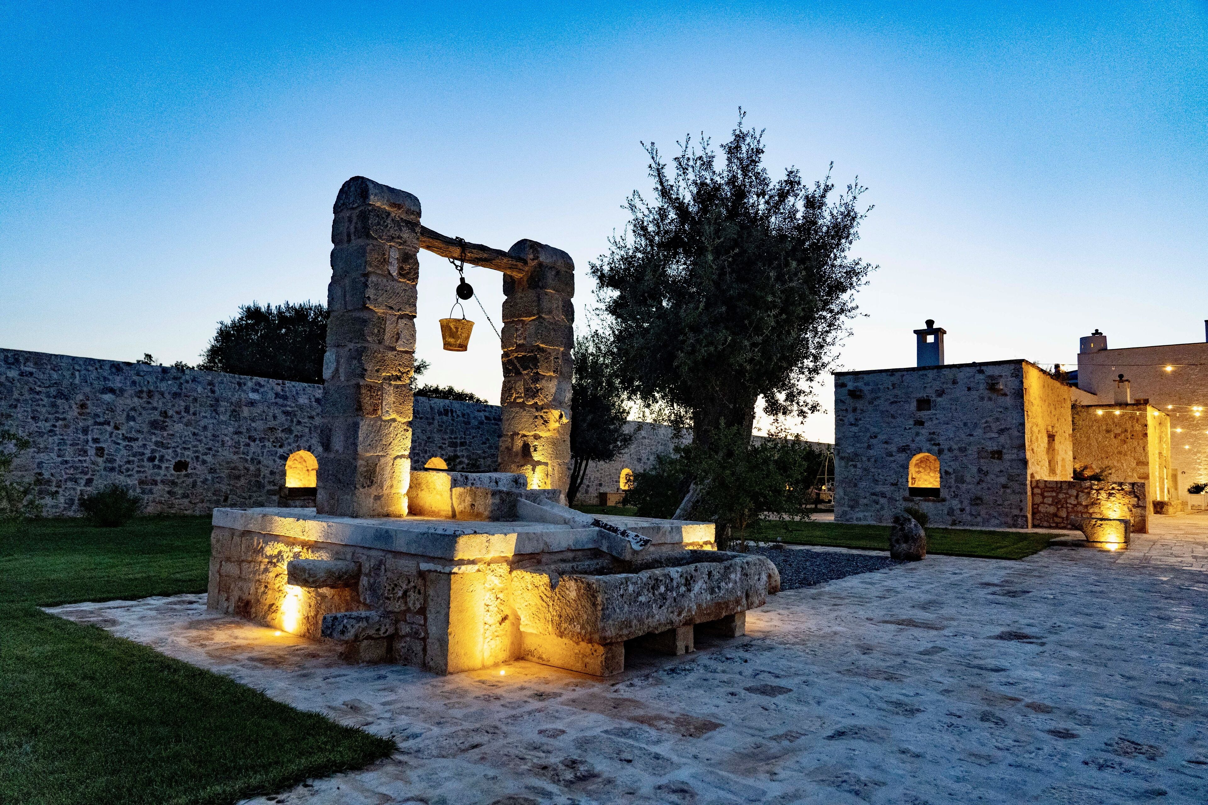 An ancient stone structure with a well-lit fountain, surrounded by a stone wall and a tree.