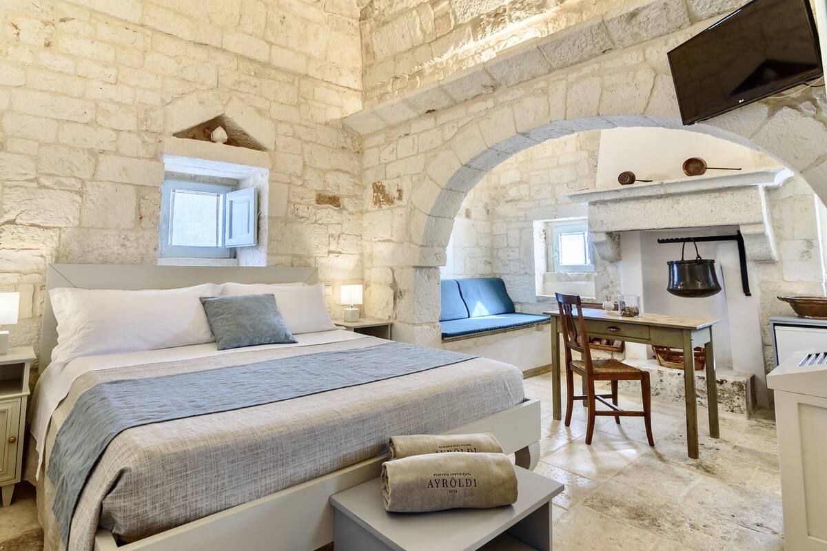 A bedroom with a stone wall, a bed with a blue blanket, a desk, and a hanging pot.