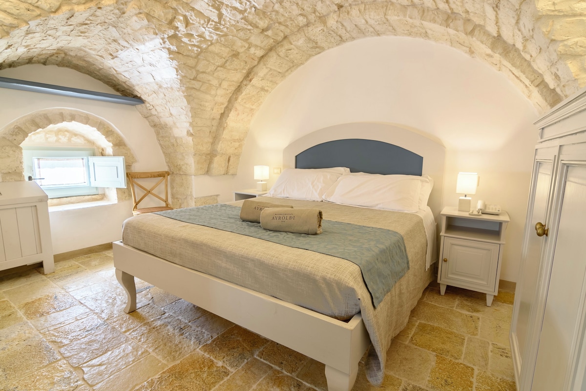 Un dormitorio con techo abovedado de piedra, una cama con cabecera color beige, una mesita de noche con lámpara y una ventana con vistas.