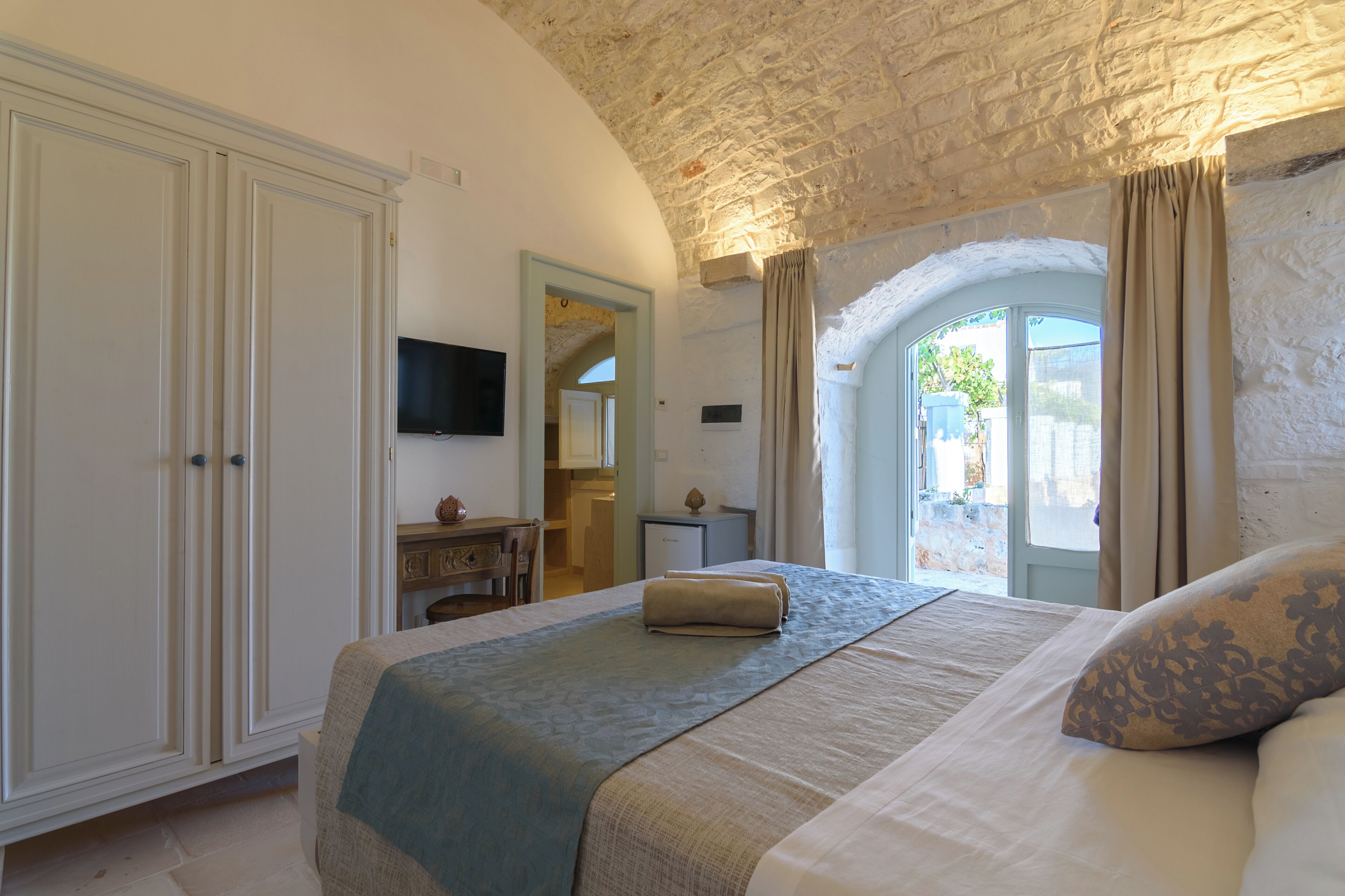 A bedroom with a bed, a wardrobe, a desk, a television, and an arched window.