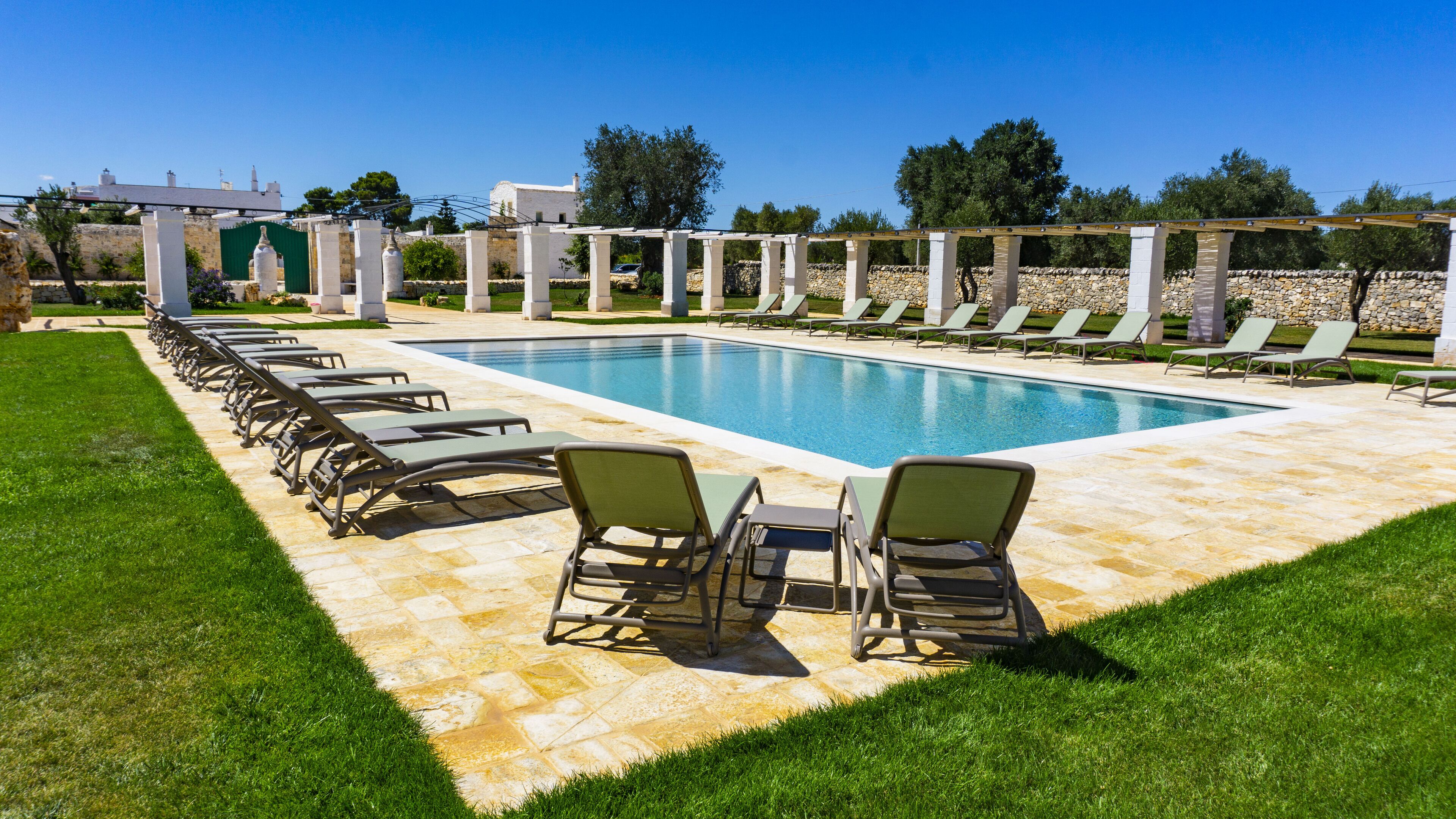 A rectangular swimming pool with a tiled deck, surrounded by lounge chairs and a pergola.