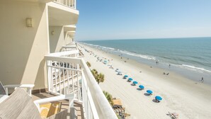 A beach view from a balcony with a clear sky, a sandy beach with umbrellas, and the ocean.