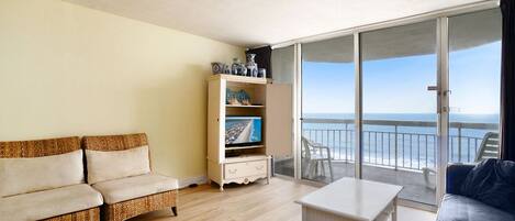 A room with a wicker sofa, a white table, a TV stand with decorative items, and a balcony with a view of the sea.