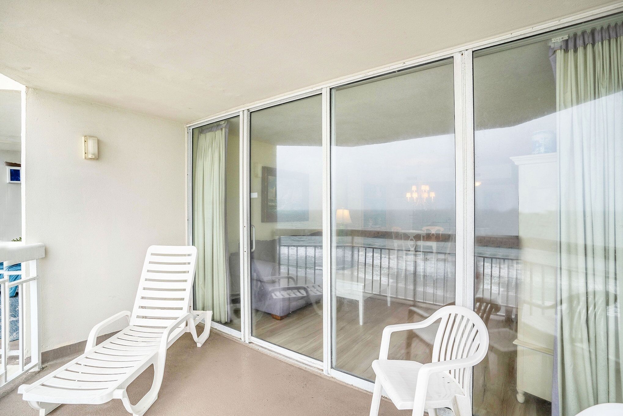 Ein Balkon mit zwei weißen Liegestühlen, einer Glastür, die auf eine Terrasse führt, und einem kleinen Tisch mit einem Stuhl.