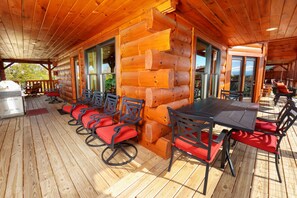 Una terraza de madera con una mesa larga y sillas, una parrilla y vistas a las montañas.
