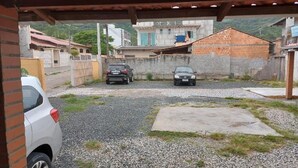 A covered parking area with two cars parked, gravel ground, and buildings in the background. - Pousada Praia do Trapiche (Penha)