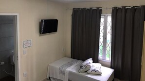 A small, single-bed room with a television mounted on the wall, a window with curtains, and a door leading to a bathroom. - Pousada Praia do Trapiche (Penha)