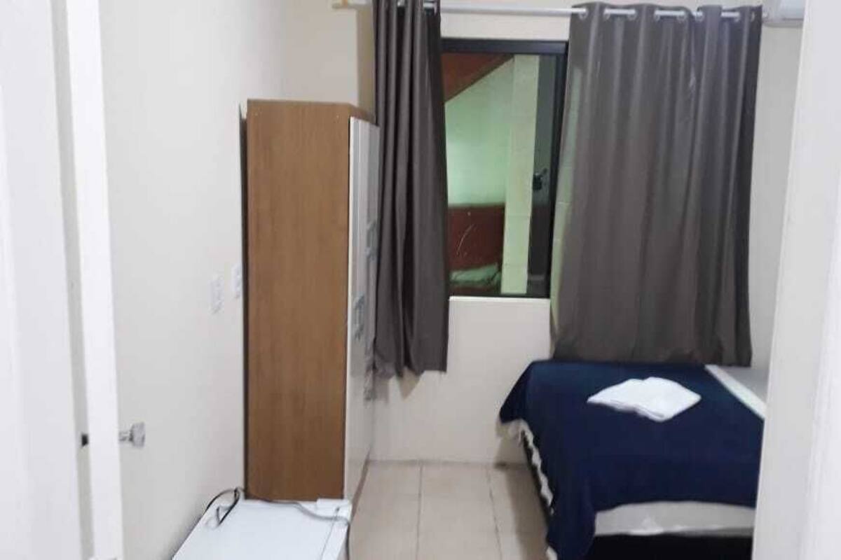 Ein kleines, schlichtes Zimmer mit einem Bett, einem Holzschrank und einem Fenster mit Vorhängen.