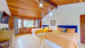 Um quarto espaçoso com duas camas, piso de madeira, ventilador de teto, televisão e janela com cortinas.