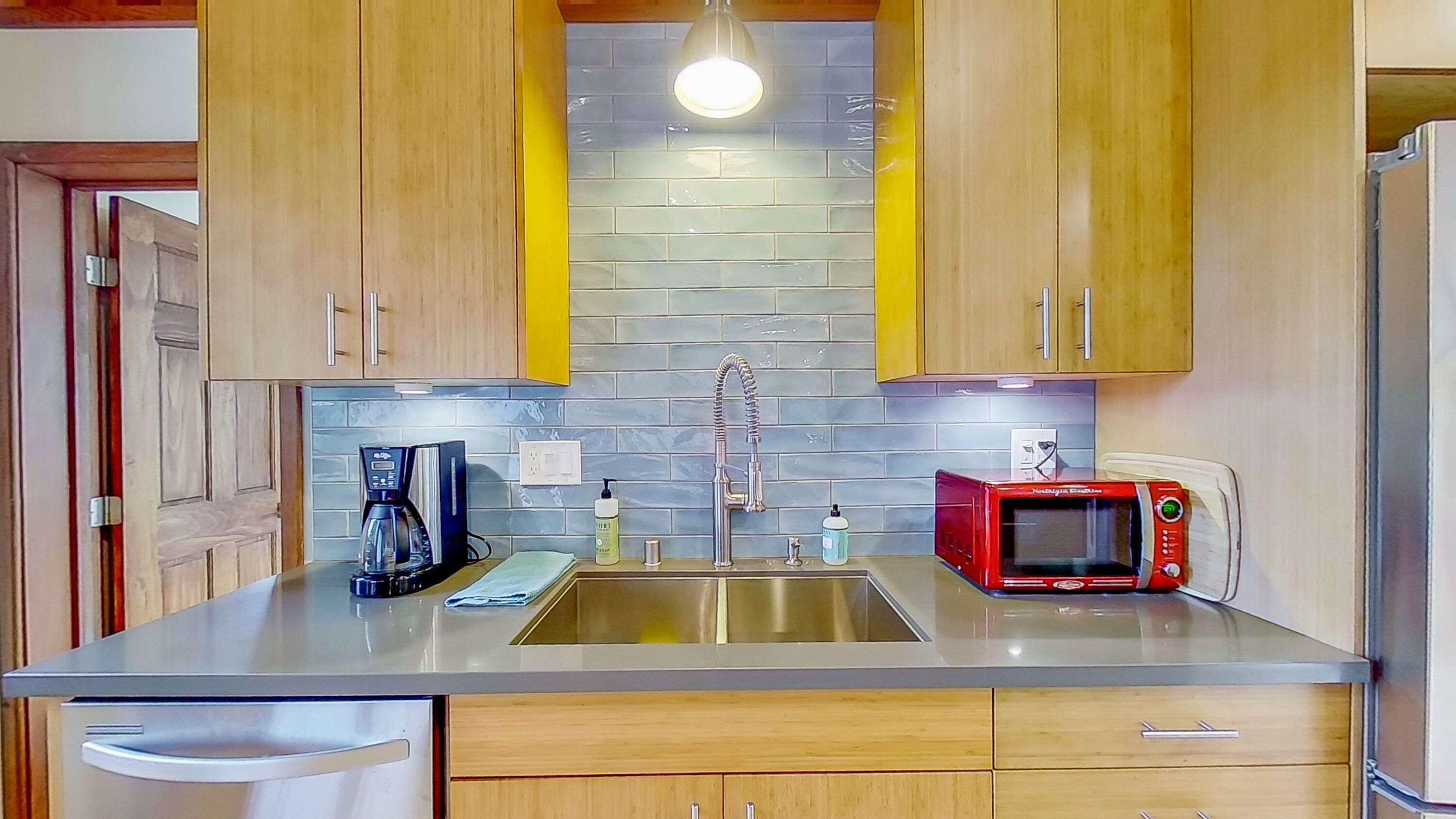 A kitchen with wooden cabinets, a stainless steel sink, a coffee maker, a microwave, and a light fixture above the sink.