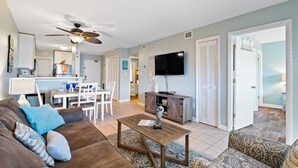 A living room with a brown sofa, patterned armchair, and a coffee table. There is a dining area with a white table and chairs, and a kitchen area with cabinets and a refrigerator. A ceiling fan is visible, and a TV is mounted on the wall. - August Specials! 3 night min! 2bed2 Bath-Walk to Beach-Indoor Pool! (Hilton Head Island)