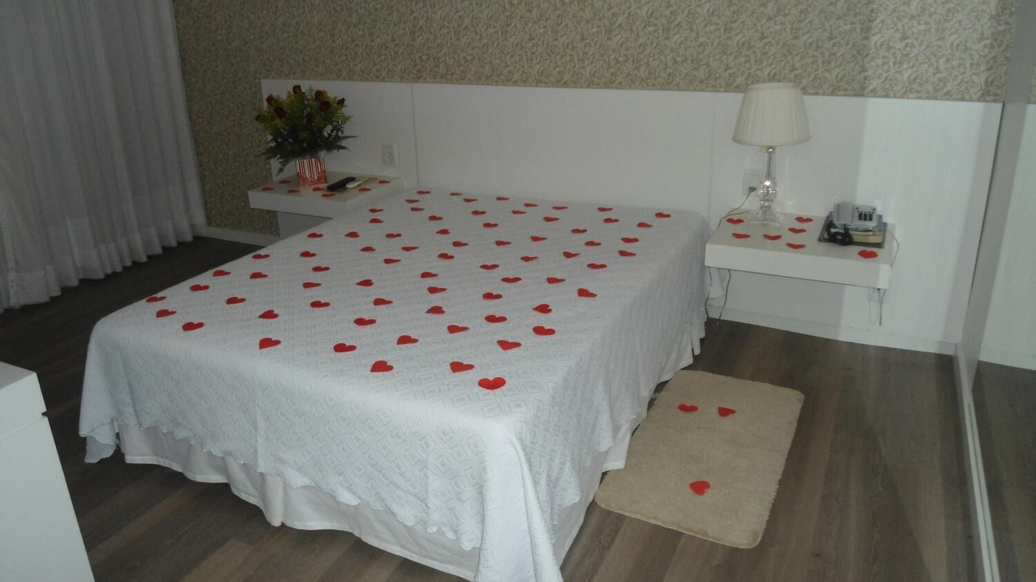 A neatly arranged bedroom with a bed covered in a white bedspread and red heart decorations, a bedside table with a lamp, and a small bedside plant.