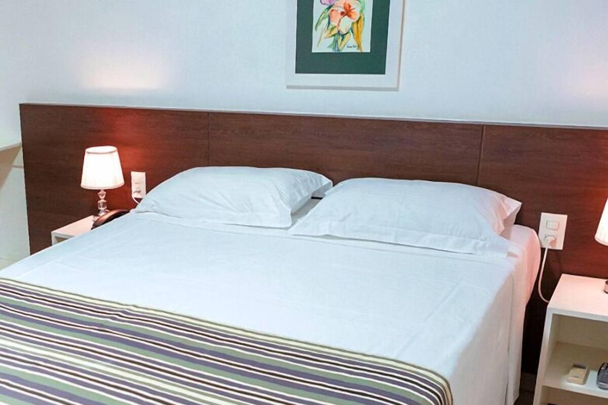 A neatly made bed with white linens, a striped blanket, and a wooden headboard. There are two bedside tables with lamps and a small shelf with a book.