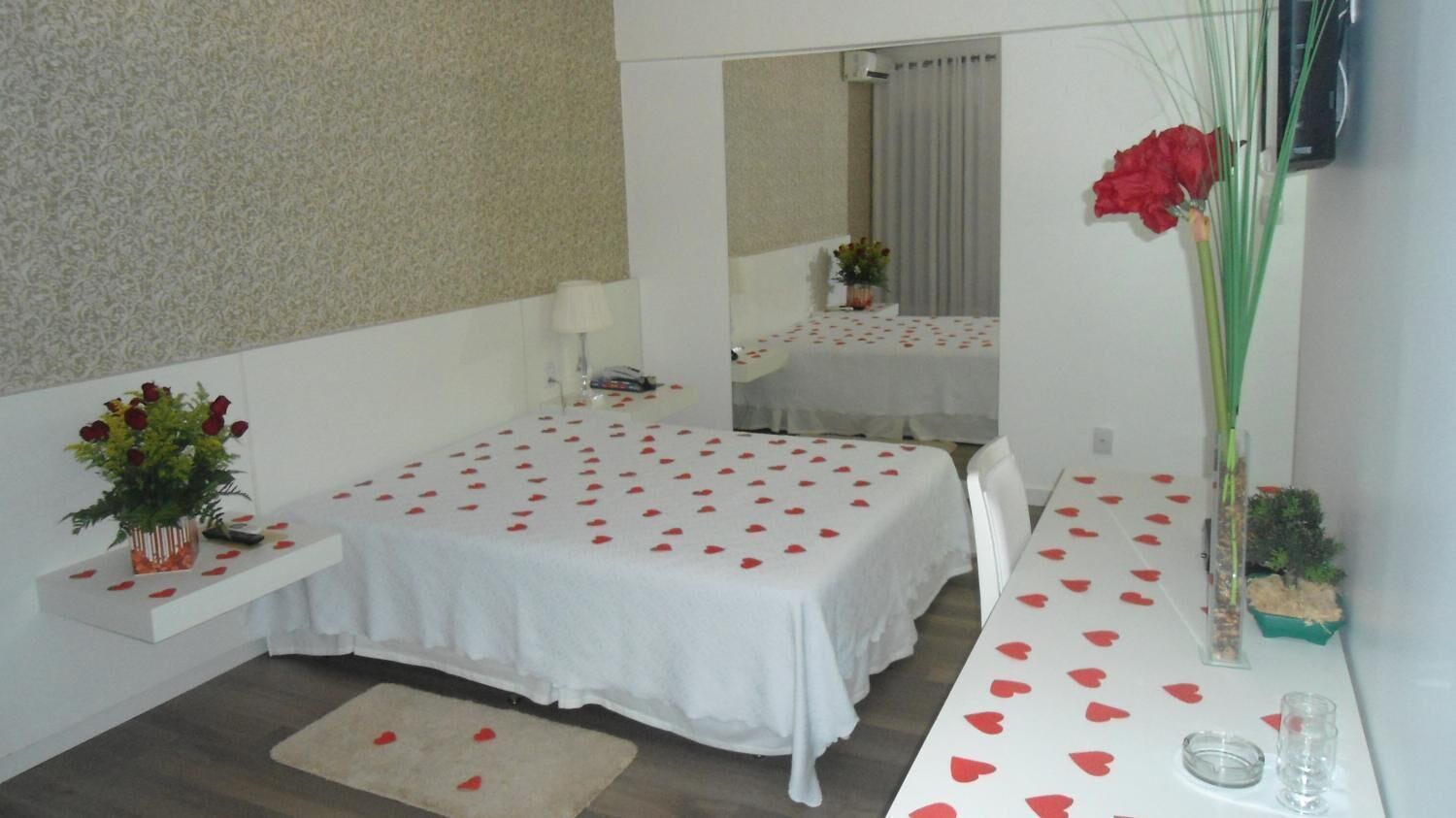 A hotel room with a bed, a small table, a mirror, and a vase with flowers.