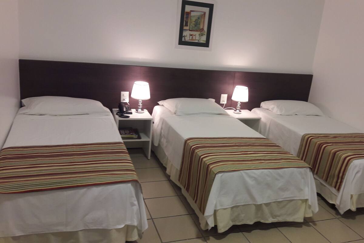 A hotel room with three single beds, each with a striped bedspread and white linens. There are bedside tables with lamps and a telephone.