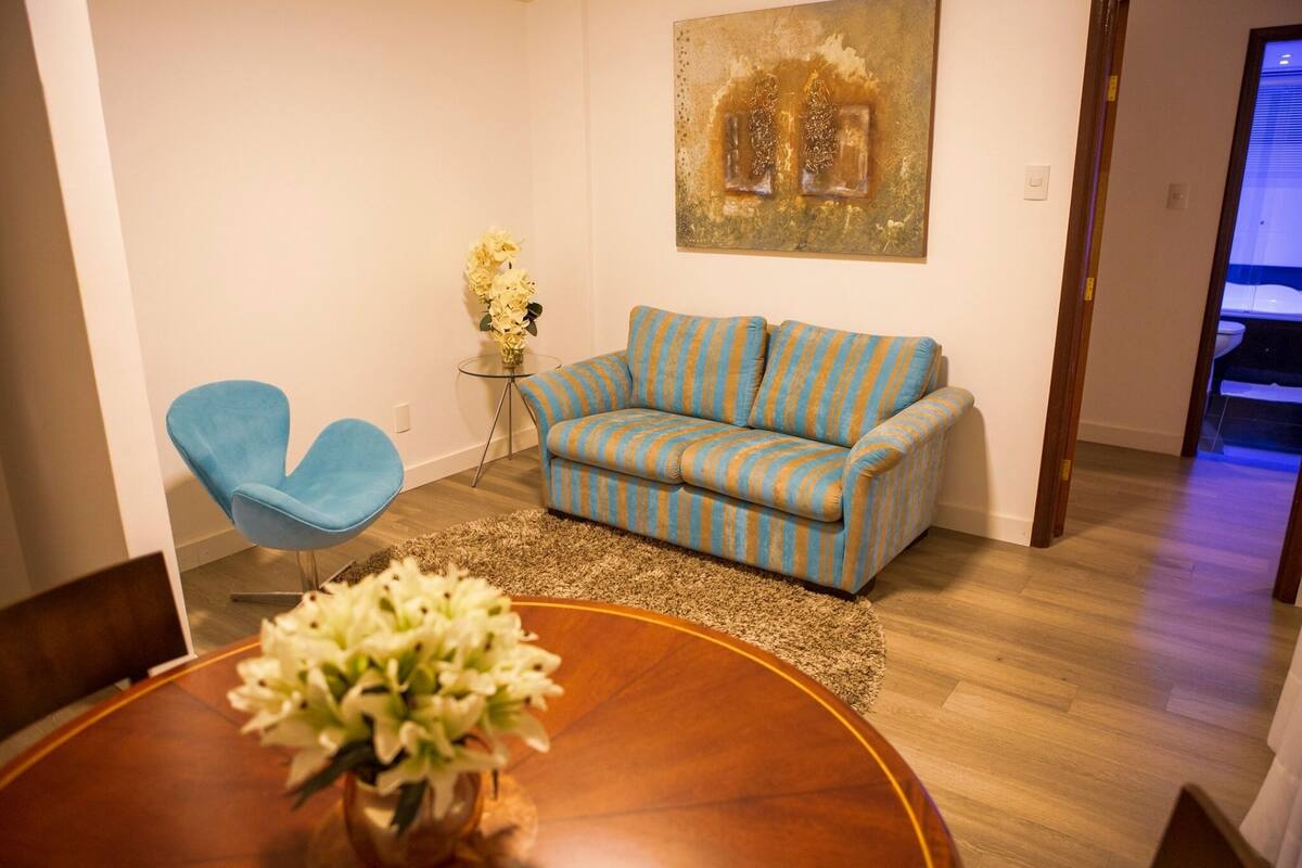 A living room with a blue armchair, a striped sofa, a round wooden coffee table with a vase of flowers, and a painting on the wall.