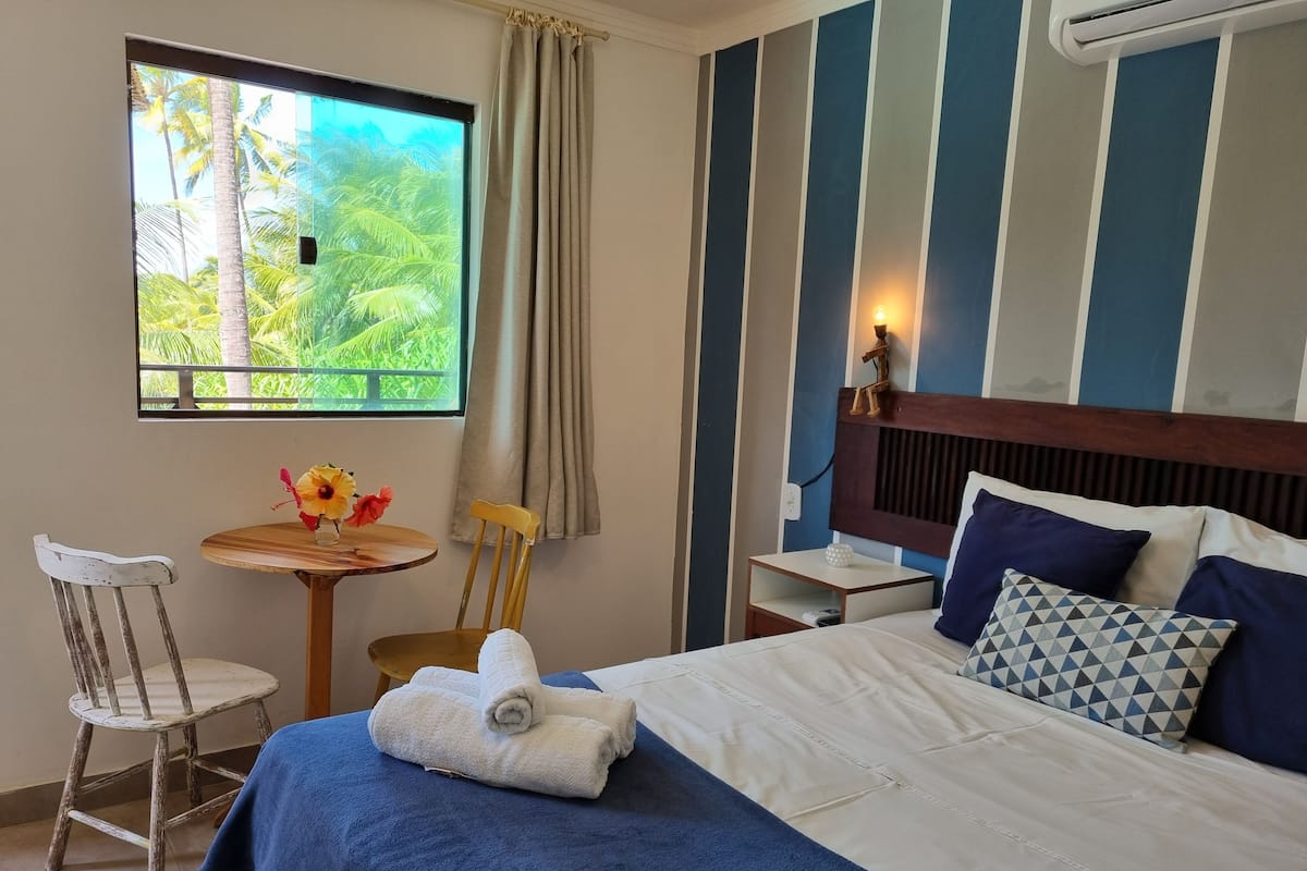 A hotel room with a bed, a small table with a vase of flowers, a chair, and a window with a view of palm trees.