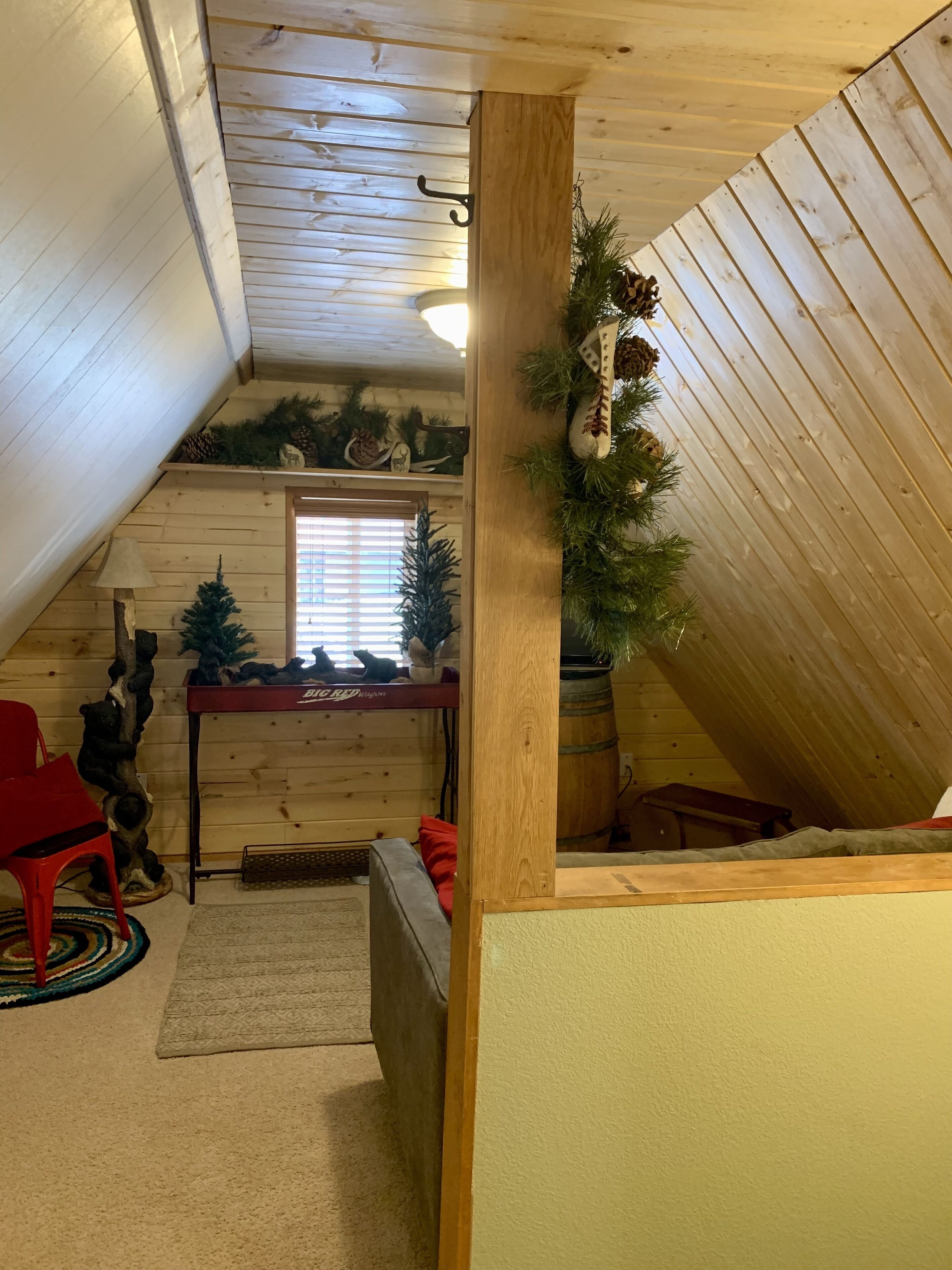 Une pièce mansardée chaleureuse avec des murs en bois, un plafond en pente et une petite fenêtre. On y trouve deux sapins de Noël, un fauteuil rouge et une table avec une lampe.