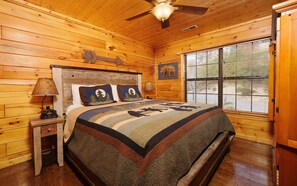 Een slaapkamer in een houten blokhut met een bed, nachtkastjes, een plafondventilator en een raam met zonwering.