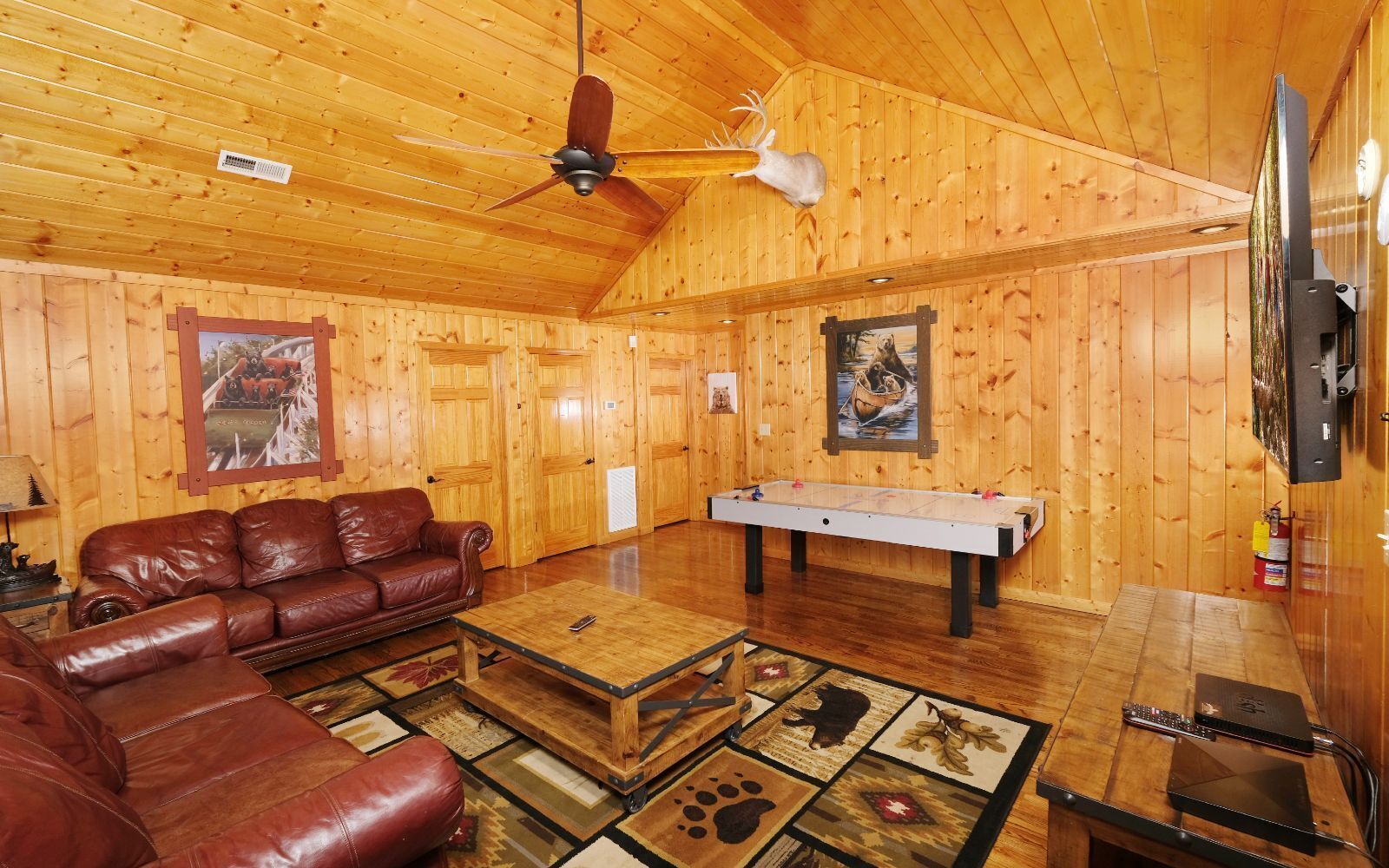 A cozy cabin interior with a leather sofa, a ping pong table, wooden walls, and a ceiling fan.