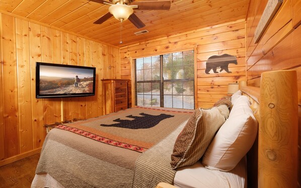 Un dormitorio de cabaña de madera con una cama, un televisor y una ventana con persianas.
