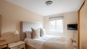 Ein Schlafzimmer mit einem Bett, einem Nachttisch mit Lampe, einem an der Wand befestigten Fernseher und einem Fenster mit Jalousien.