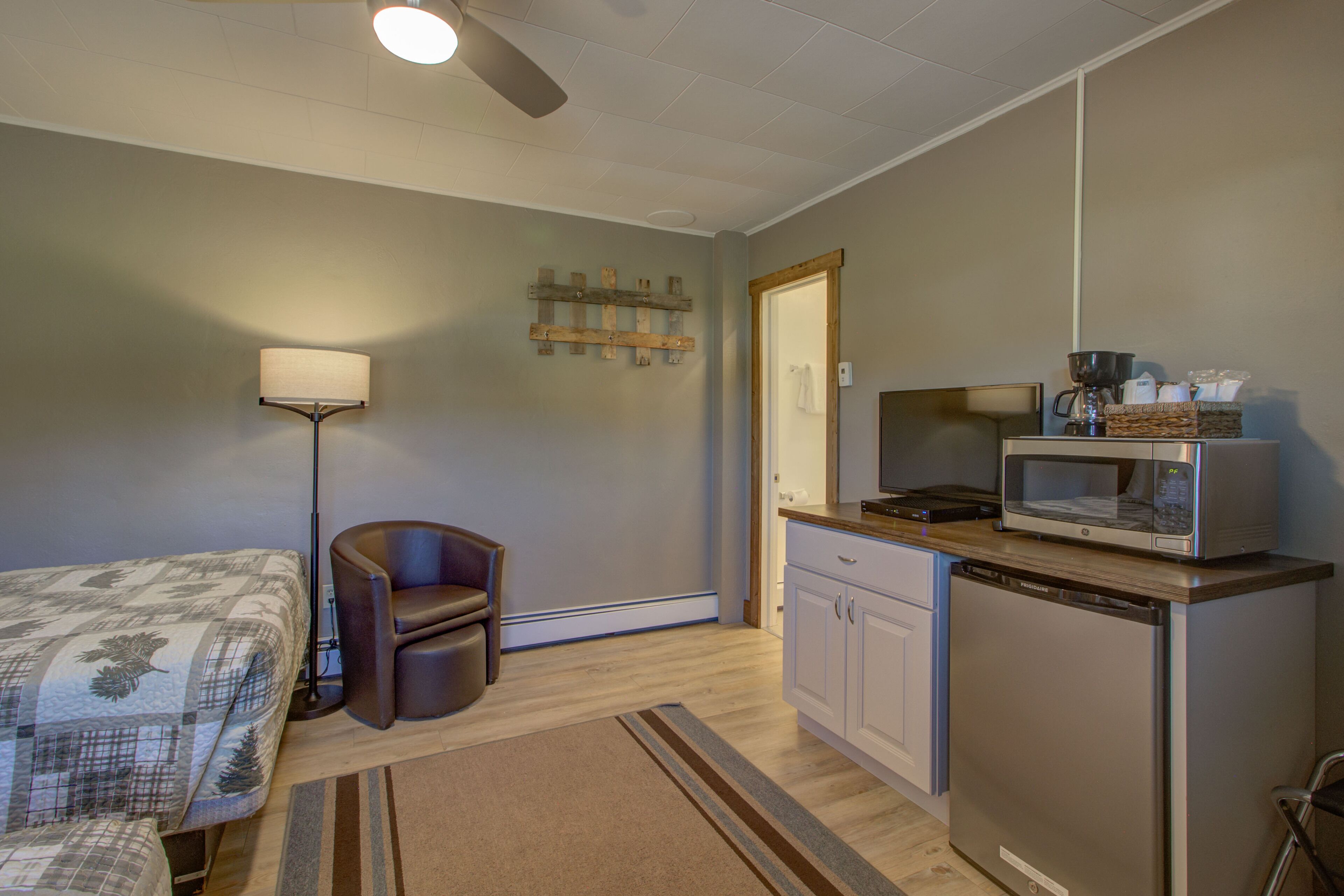 A compact room with a bed, a chair, a kitchenette with a microwave, and a ceiling fan.