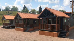 Een rij houten chalets met metalen daken, gelegen op een grindpad, met op de achtergrond bomen en heuvels.