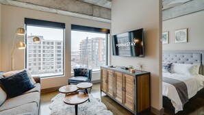 A modern hotel room with a large window, a flat-screen TV, a tufted headboard, and a wooden cabinet. - MINNeSTAY* Sable 507 | North Loop | Target Center (Minneapolis)