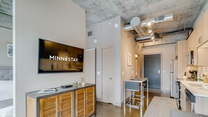 A modern hotel room with a large flat-screen TV, wooden cabinets, a kitchenette, and a dining area. - MINNeSTAY* Sable 507 | North Loop | Target Center (Minneapolis)