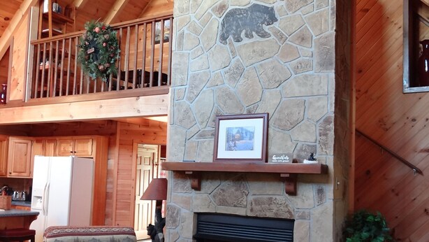 Un intérieur de chalet chaleureux avec une cheminée en pierre, des poutres en bois, un balcon décoré d’une guirlande et un fauteuil sur le thème de l’amour.