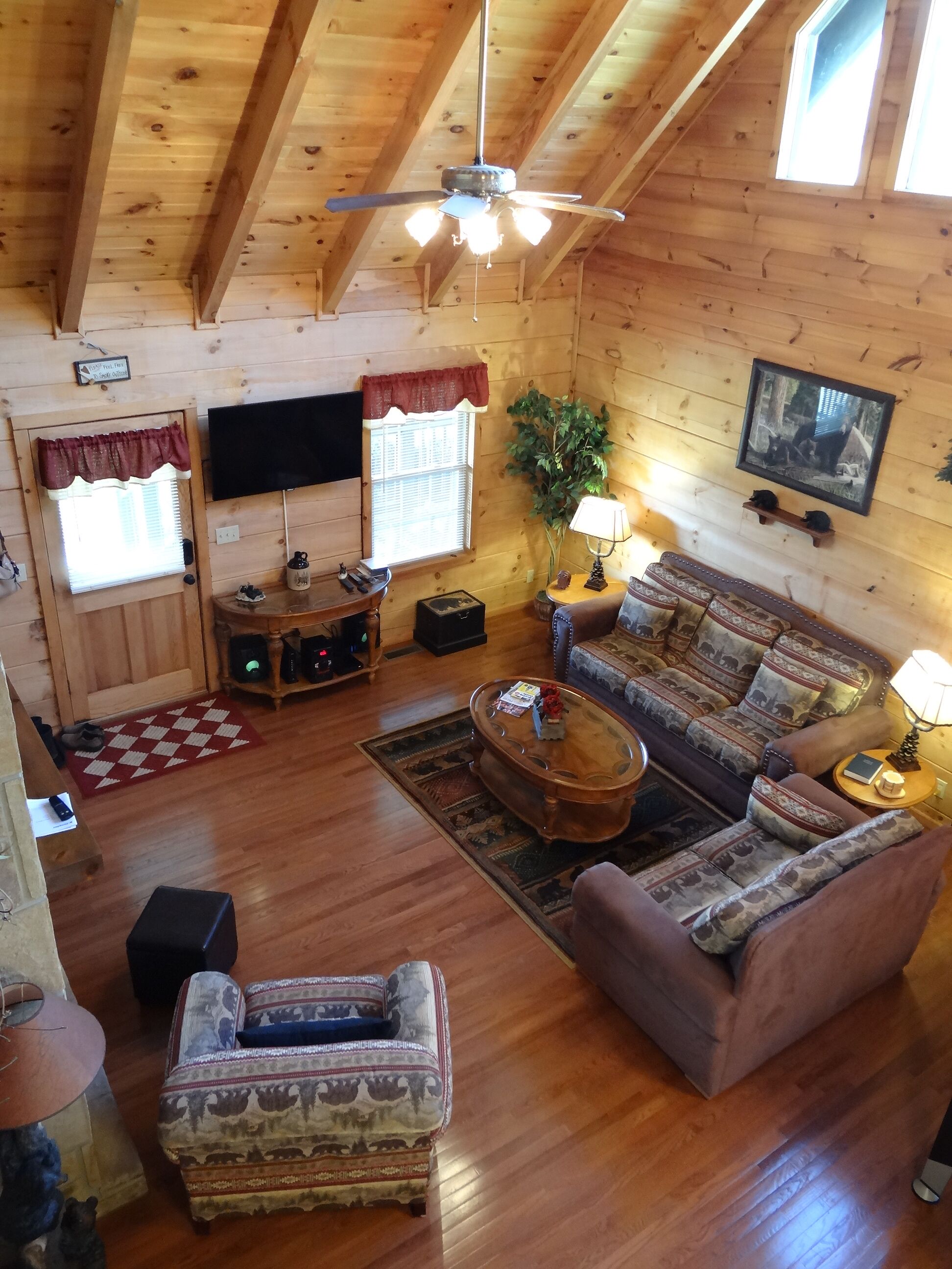 A cozy log cabin interior with a wooden ceiling, a ceiling fan, a television, a sofa, a coffee table, and a patterned rug.