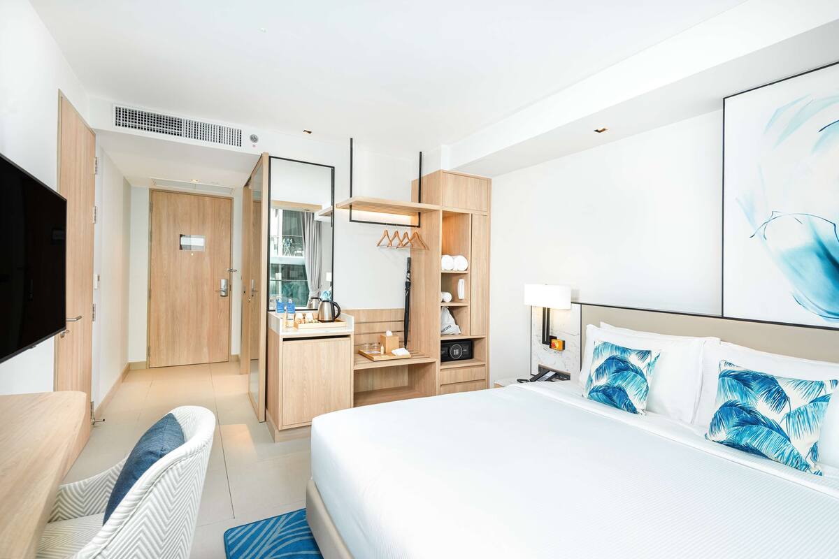 A modern hotel room with a large bed, a sitting area with a chair, a kitchenette, a wardrobe, and a flat-screen TV.