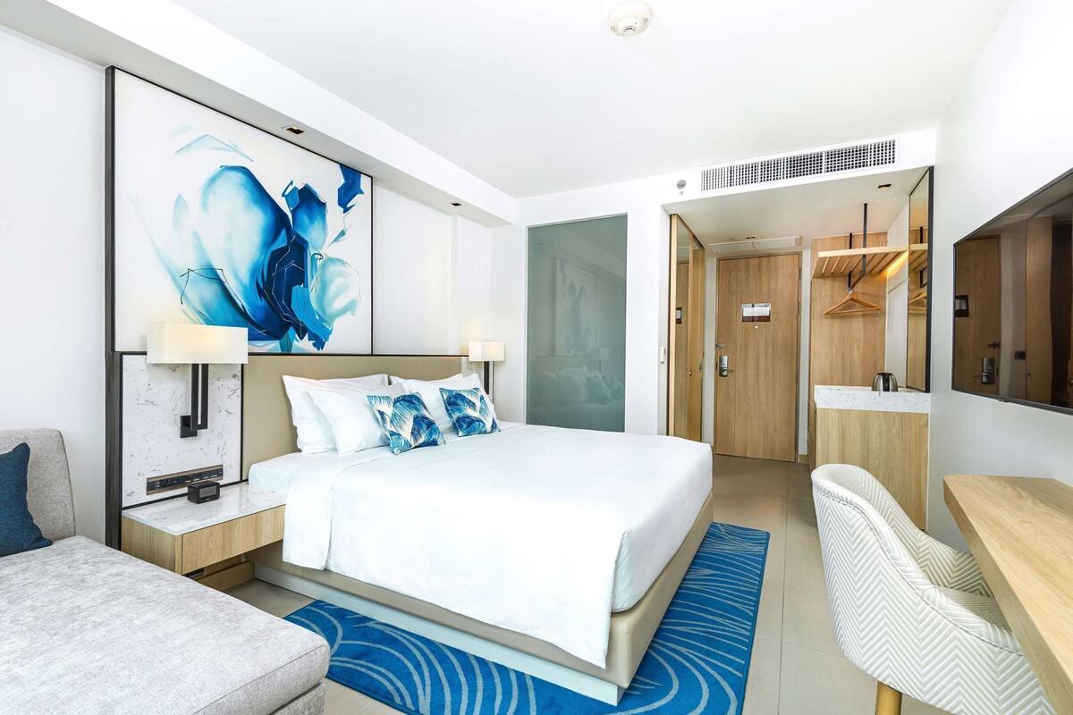 A modern hotel room with a large bed, a desk, a chair, a wardrobe, and a bathroom visible through an open door.